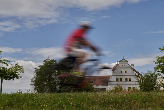 Radfahrer in einem roten T-Shirt auf einem Fahrradweg. Im Hintergrund sind weiß gestrichene Häuser zu sehen. Im Vordergrund eine Wiese.